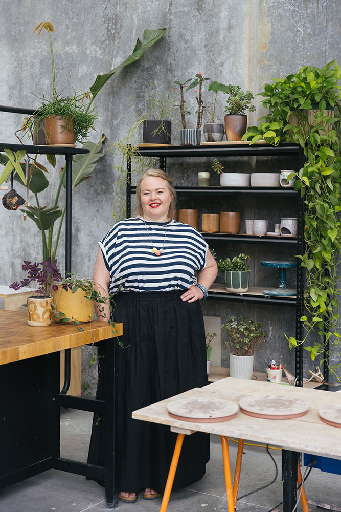 Ceramicist Arcadia Scott stands amongst her studio space, surrounded by shelves of her handmade work and indoor plants