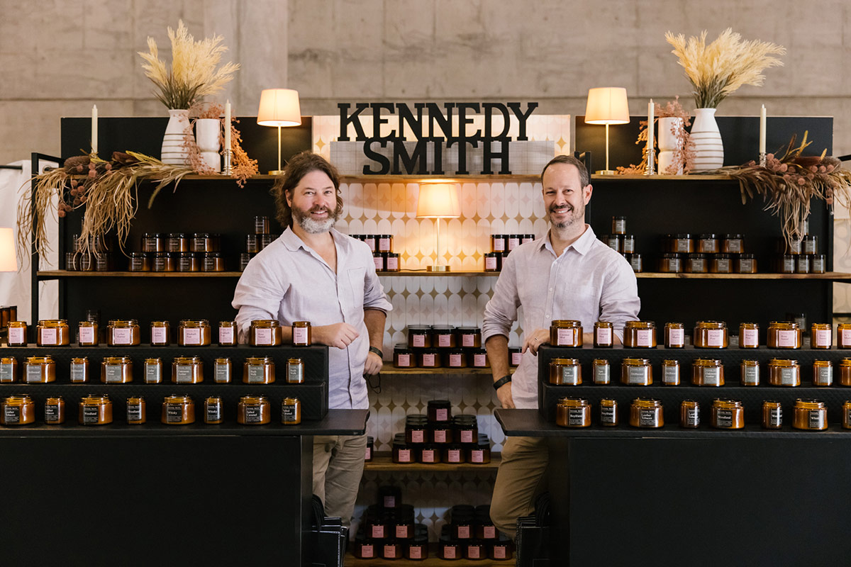Portrait of the founders of Kenneth Smith candles pose in front of their boutique-like market stand display