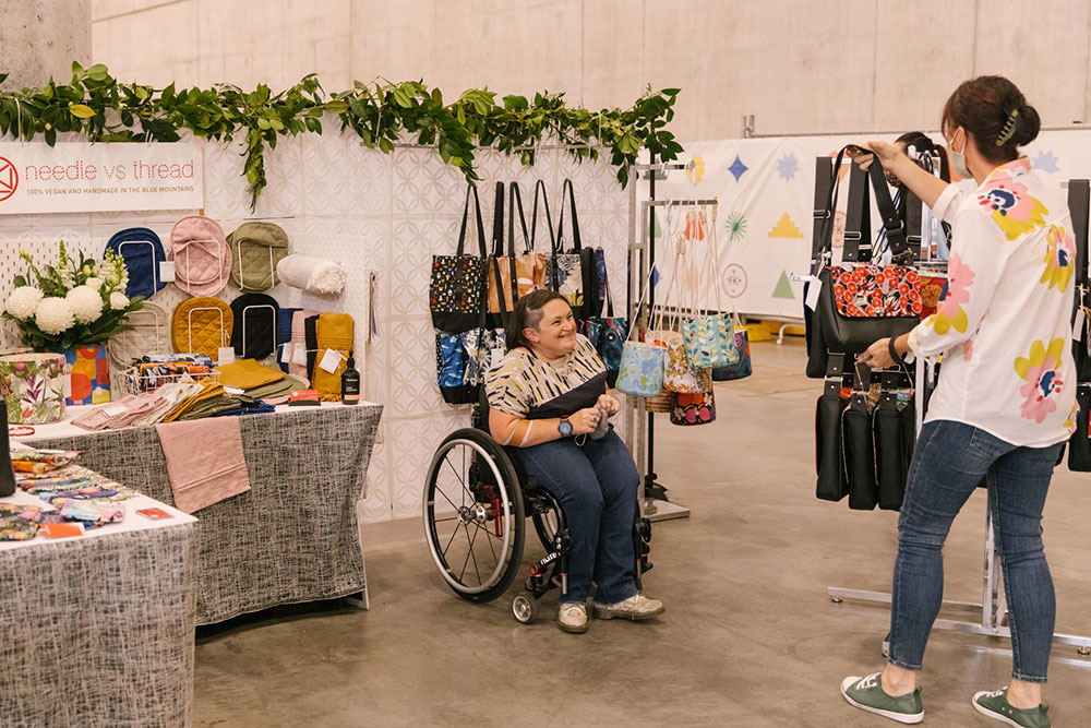 A woman in a wheelchair chats about their designs with a customer at their market stall
