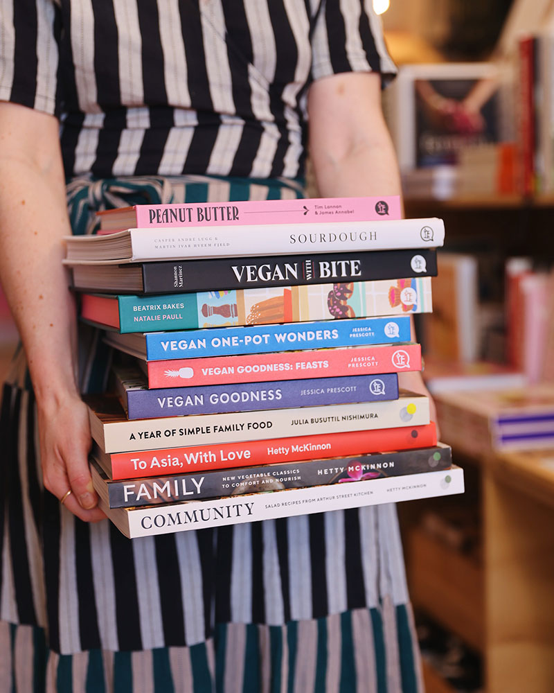 Woman holds a stack of colourful cookbooks in front of her in the shop A Quirk of Fate Northcote Melbourne
