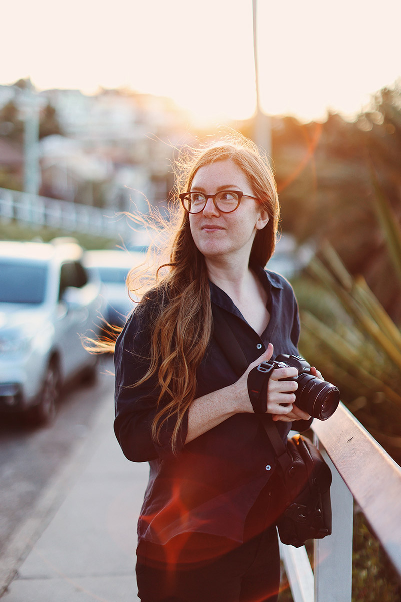 Portrait of Samee Lapham standing on the street at sunset with her camera in her hand and sun behind her, illuminating her long strawberry blonde hair in the breeze. She is wearing glasses, dressed in a navy linen shirt, and is smiling with her mouth closed and her eyes look off to the left.