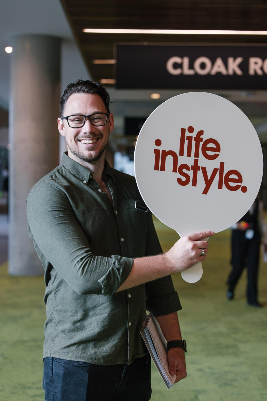 A male smiles at the camera holding a sign with the Life Instyle logo as a form of direction to arriving attendees
