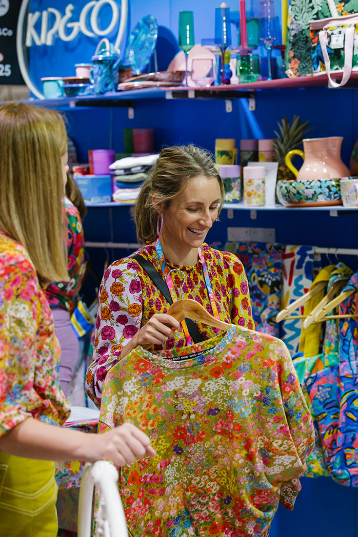Portrait of smiling female retailer at stall by Kip and Co holding a colourfully printed garment and surrounded by products on display