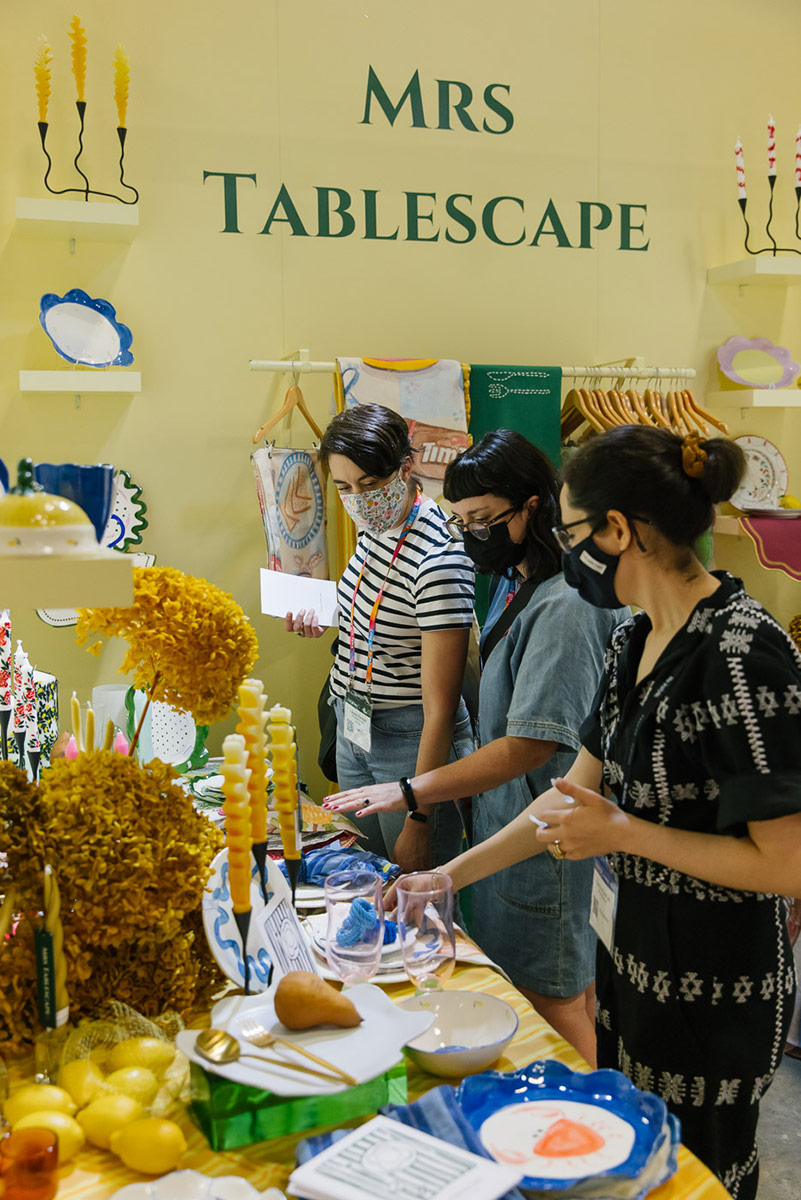 Three women browse and discuss homewares products at the Mrs Tablescape stand, wearing masks