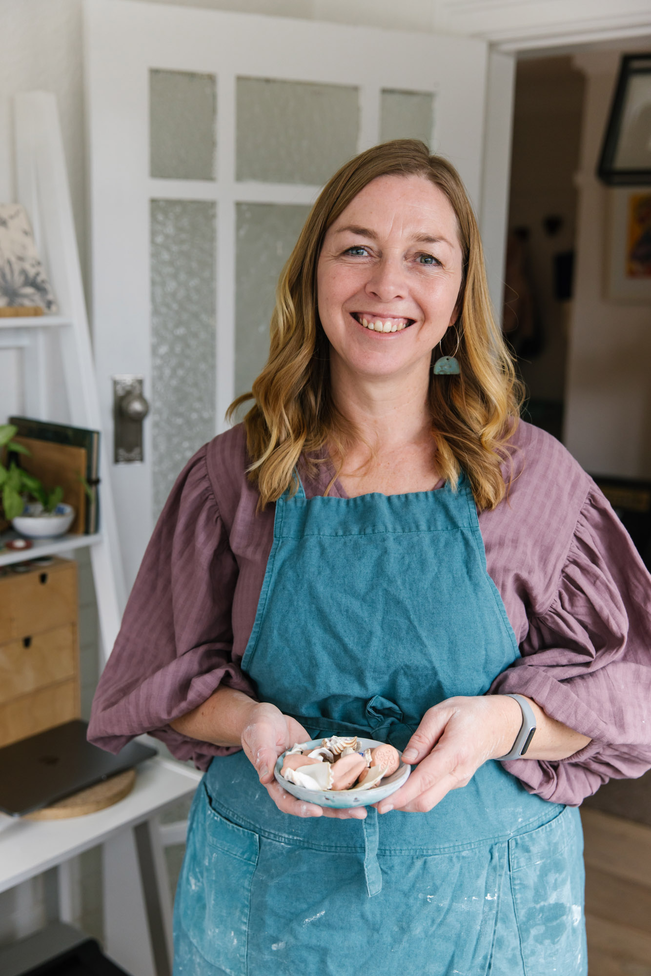 Portrait of Meraki Fire Ceramics maker Nicole Redenbach wearing a blue apron and holding a plate of handmade stamps and shells