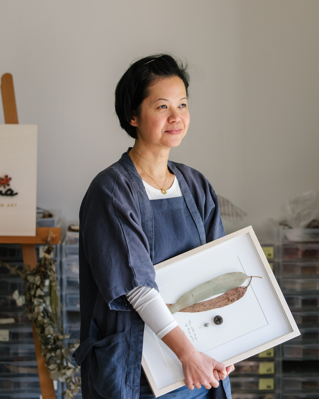 Sophie, an asian woman with short black hair, stands looking out the window holding a framed botanical artwork