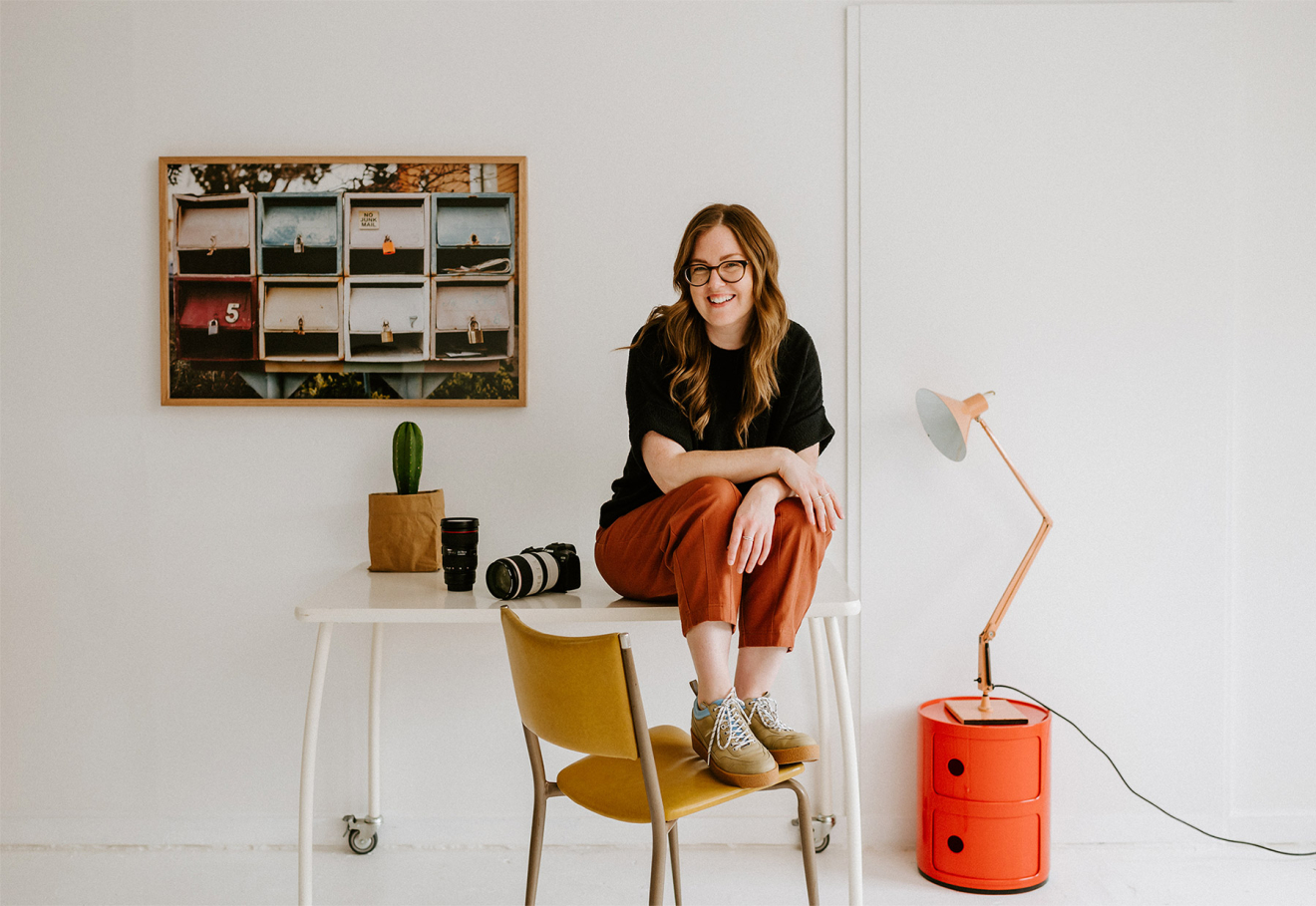 Sydney photographer Samee Lapham sits on a table in a white studio room with her feet on a mustard colour chair, am orange retro lamp to the right and a framed print on the wall to the left.