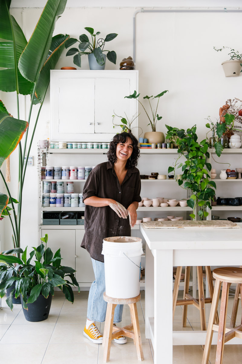 A woman stands in her ceramics studio surrounded by glazes, shelves of work and green indoor plants, while she prepares recycled clay from a white bucket