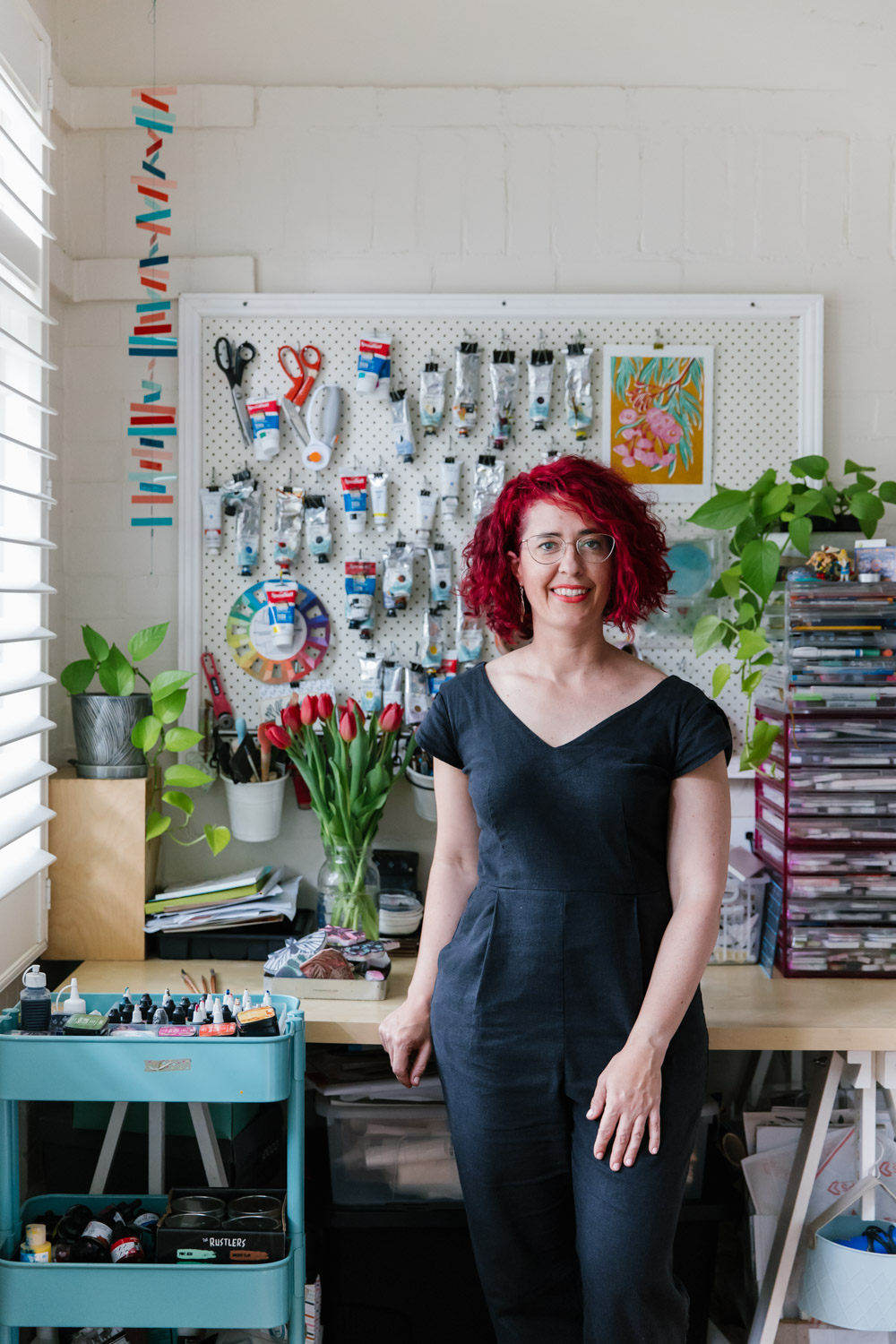 Portrait of Rowan Sivyer, a white woman with bright red hair wearing a black jumpsuit, standing in her home studio where she creates block printing artworks by hand