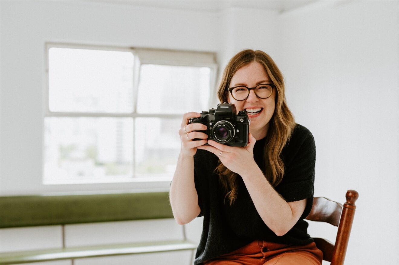 Portrait of a white woman with strawberry blonde hair and glasses holds a vintage camera and laughs