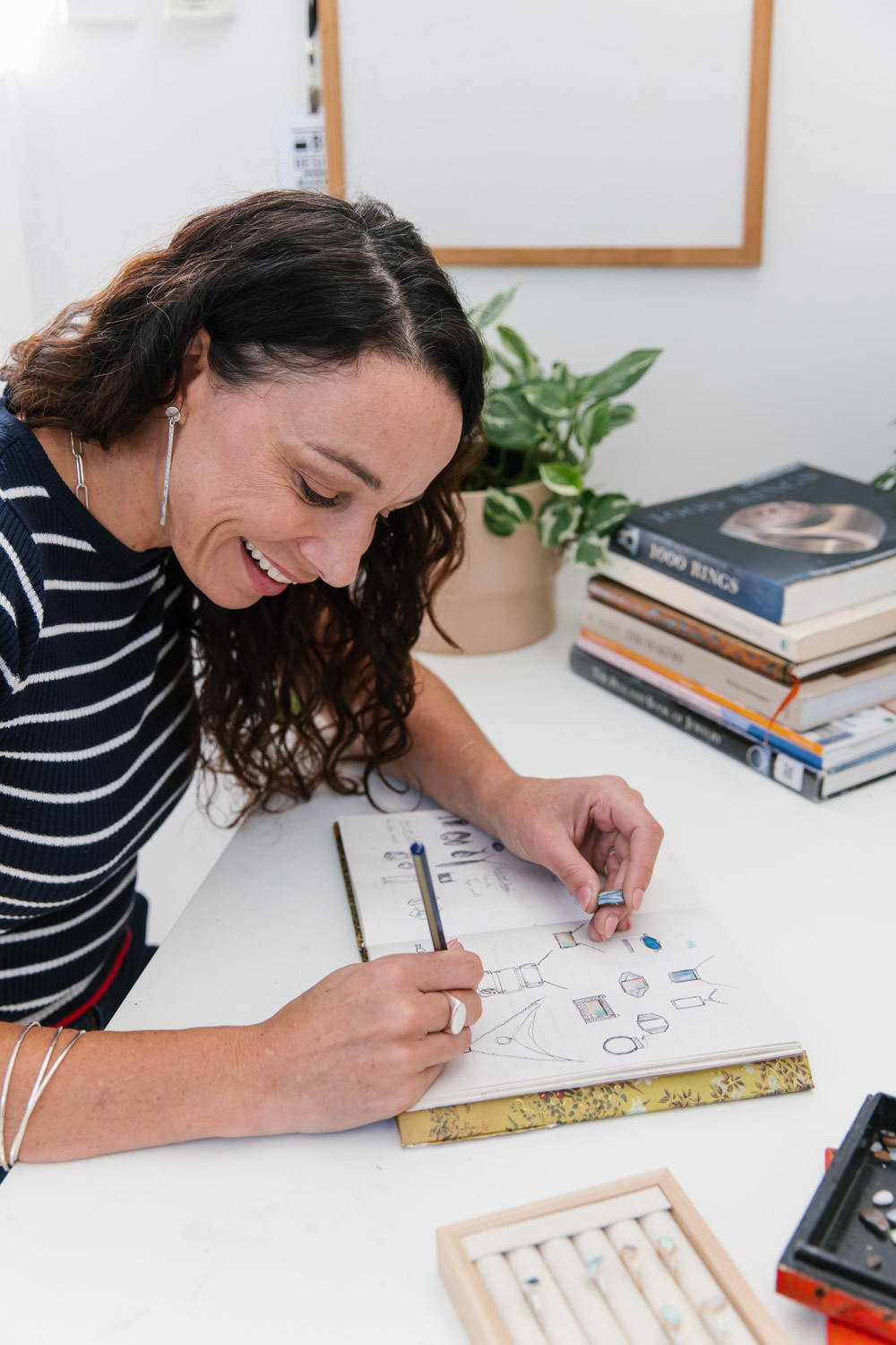 A white women with curly brown hair sits at her desk sketching jewellery design ideas