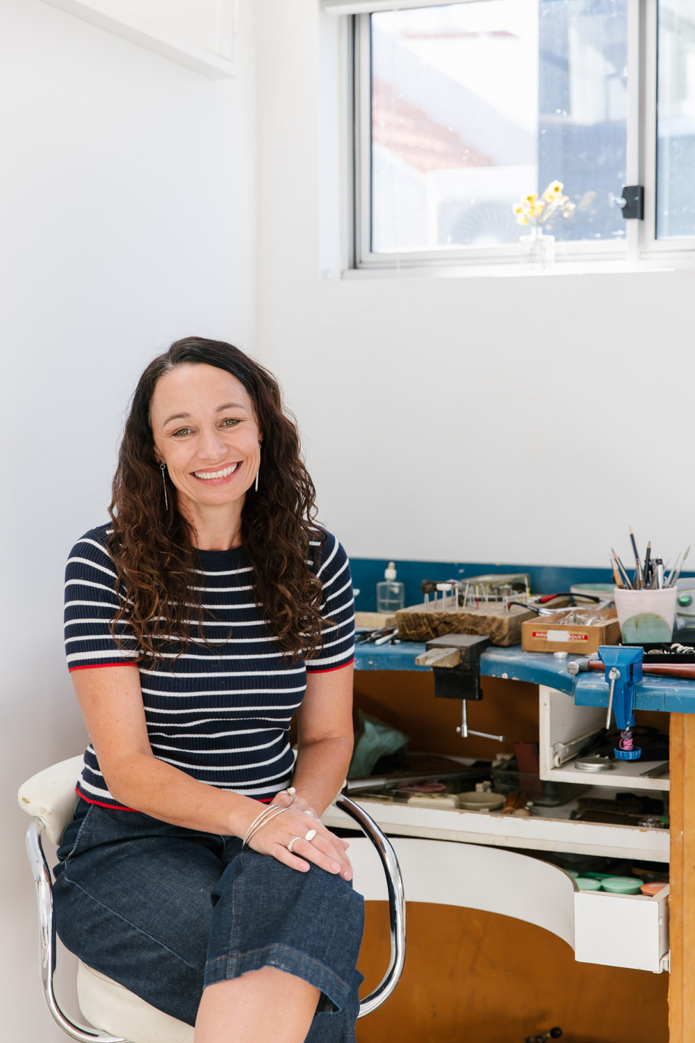 White woman with curly brown hair sits at her jewellers workbench, smiling at the camera and wearing a navy and white striped top and denim pants