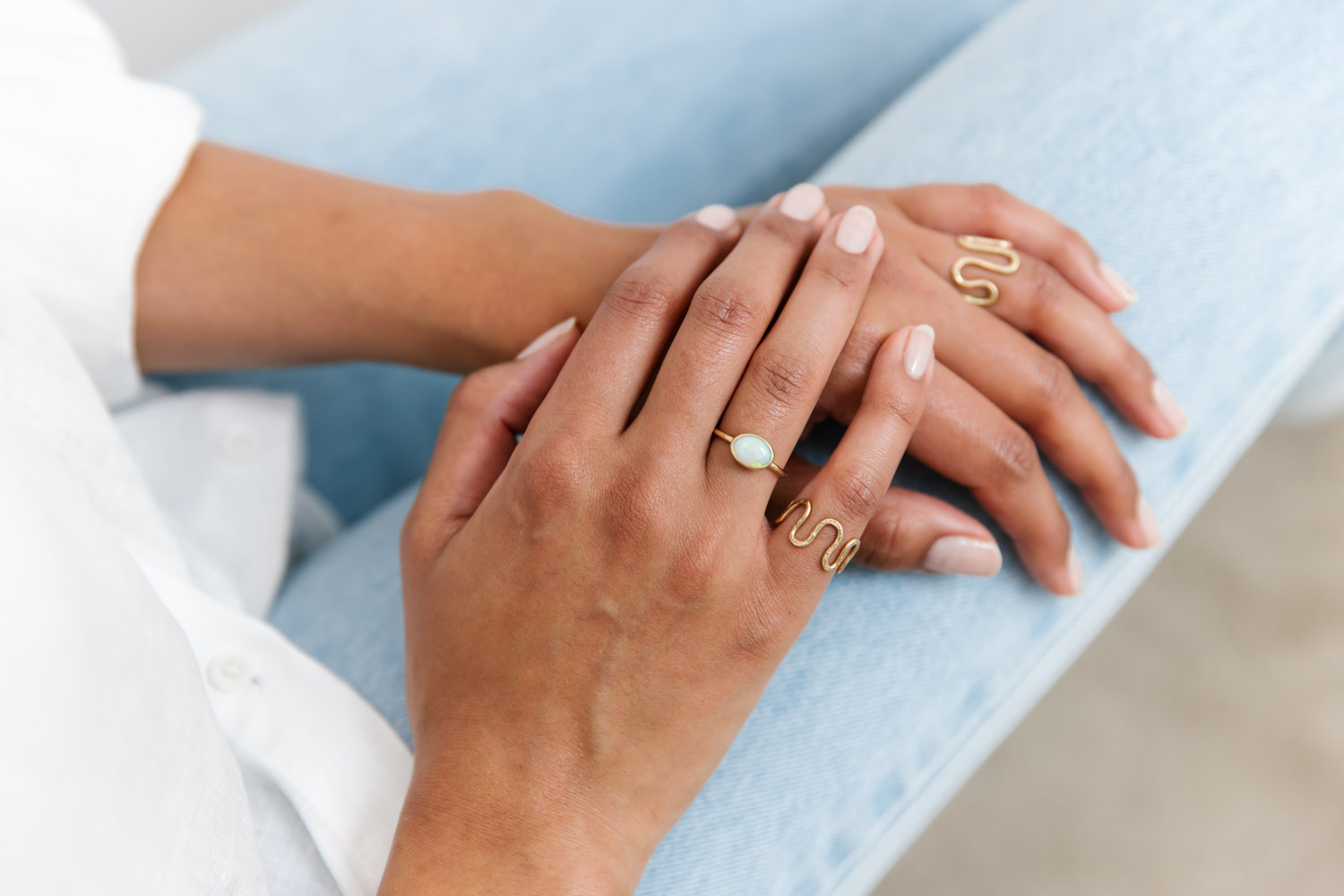 Women of colour wears rings created by Jane Jones Jewellery, with her hands resting on her leg, wearing light denim jeans and white linen top