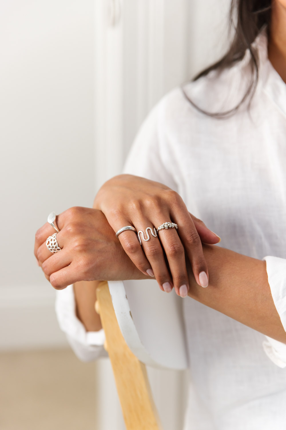Woman wears a range of sterling silver rings, resting her hands over the back of a white chair, wearing a white linen shirt