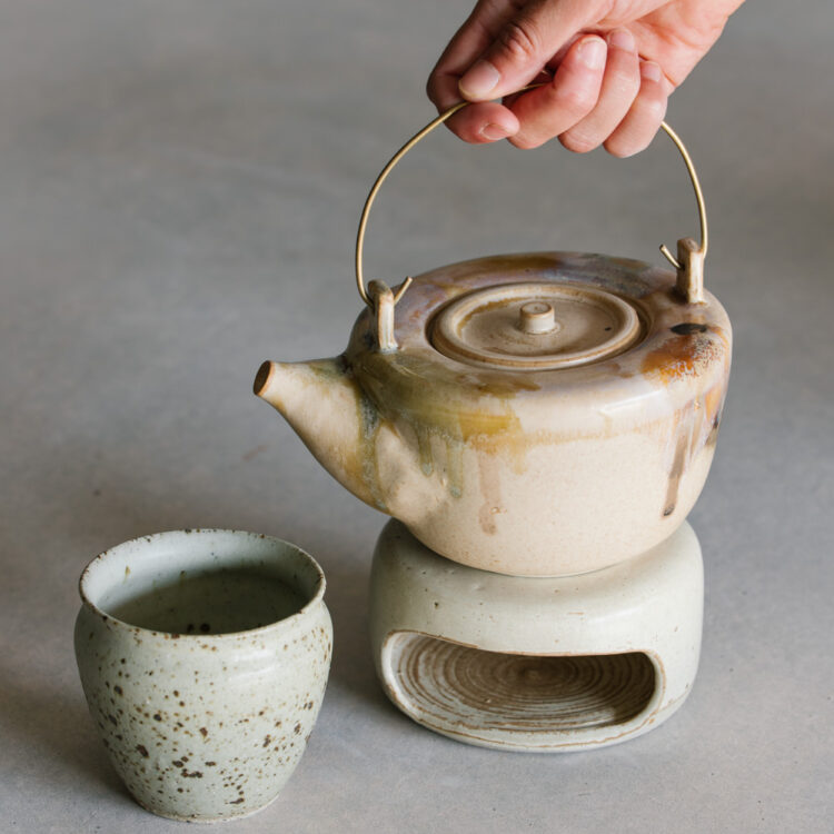 Ceramic teapot on a teapot warmer and accompanying teacup, with unique glazes dripping down the sides in neutral tones made by Emi Ceramics