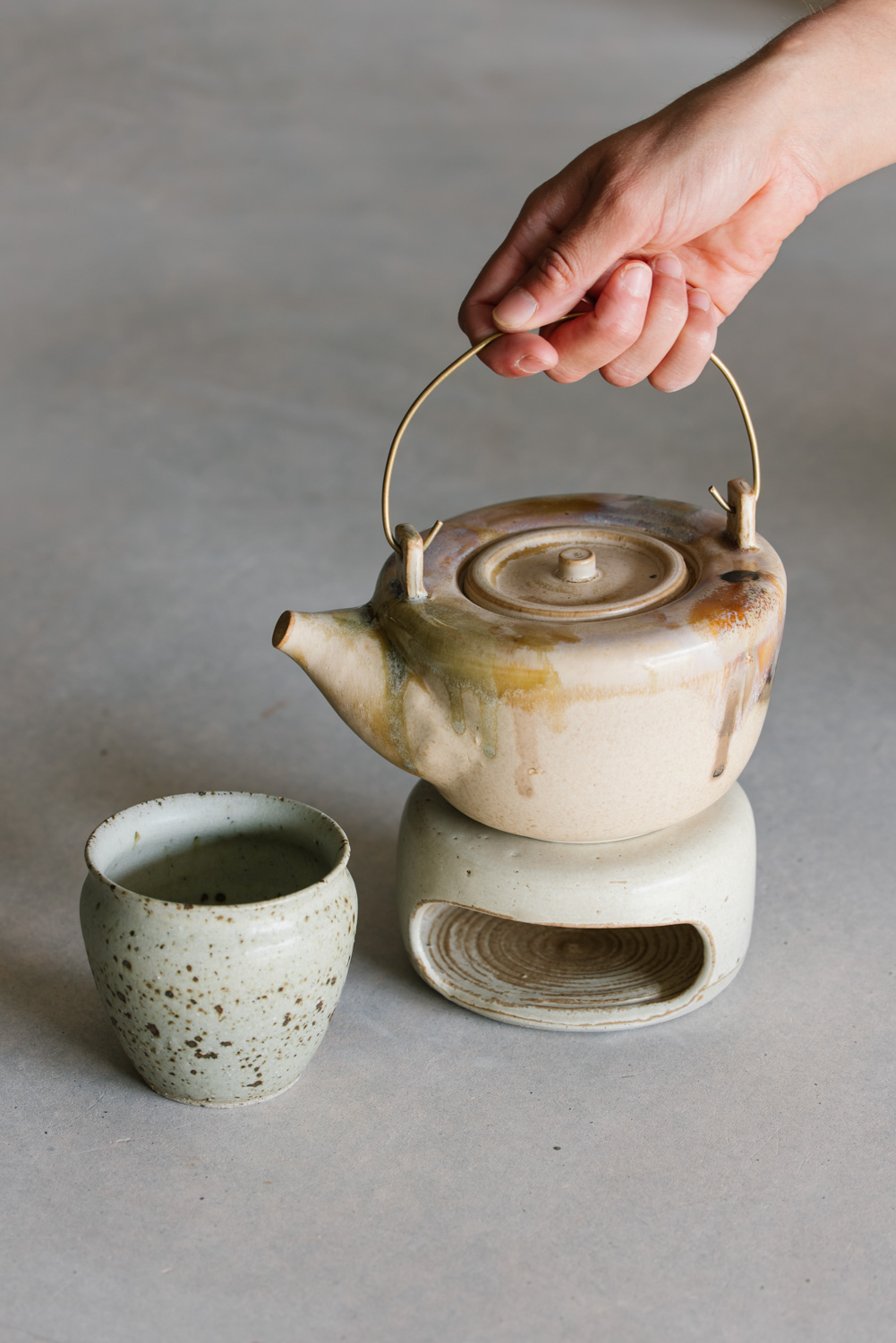 Ceramic teapot on a teapot warmer and accompanying teacup, with unique glazes dripping down the sides in neutral tones made by Emi Ceramics